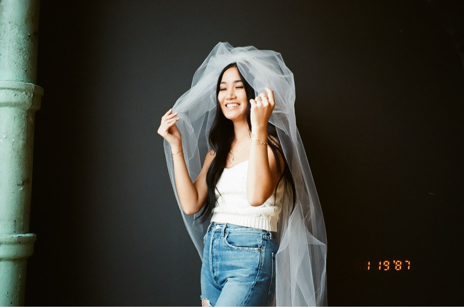 A girl standing in front of a black wall wearing a veil, a  white tank and jeans laughing. Shot on 35mm film.