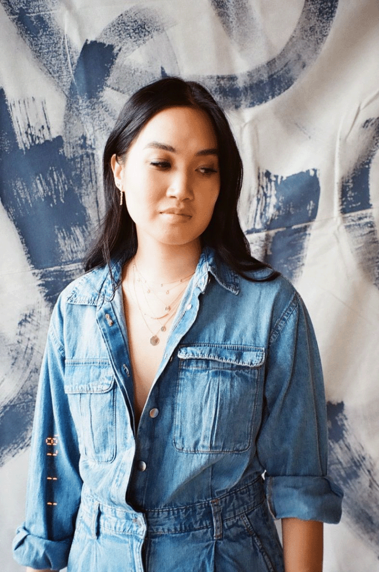 A girl standing in front of a painted backdrop wearing a blue denim jumpsuit and a necklace stack. Shot on 35mm film.