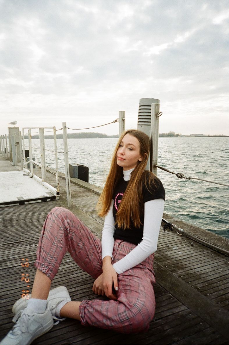 Zoe sitting on a dock in Toronto wearing pink plaid pants, a black baby tee and a white turtleneck. Shot on 35mm film.