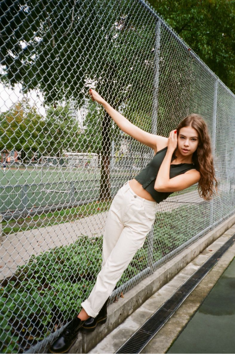 A girl holding onto a metal fence in a green tank top and white jeans. Shot on 35mm film. 
