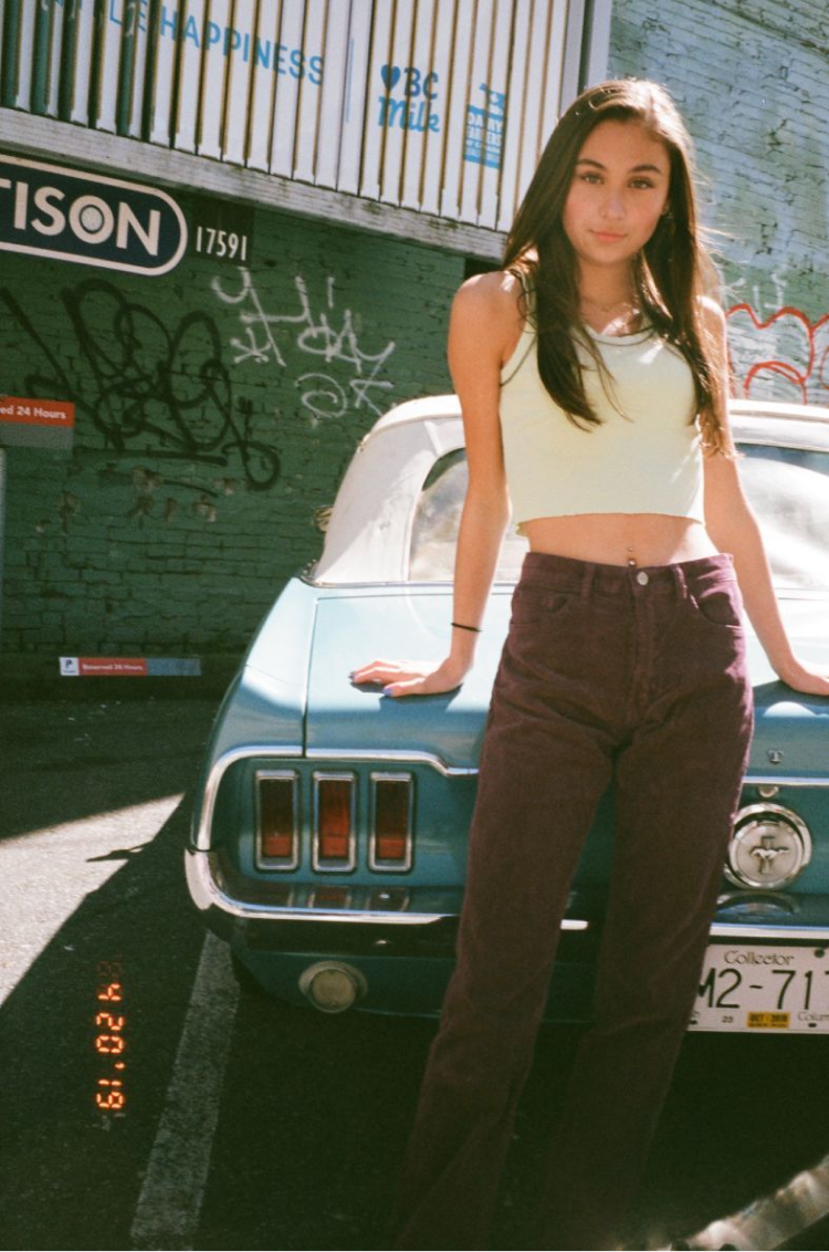 A girl in a green tank and purple corduroy pants posing in front of an antique car. Shot on 35mm film.